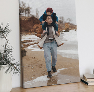 A photo canvas displayed on a wooden surface shows a man walking along a sandy beach with a child sitting on his shoulders. They are dressed in winter clothing, and waves gently wash up near their feet. A potted evergreen plant and stacked books sit nearby, adding a cozy indoor touch.