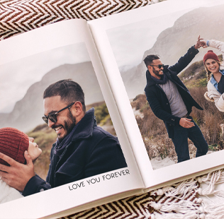 An open photo book resting on a cozy blanket shows a couple laughing and enjoying time outdoors. One photo captures the man smiling as he holds the woman’s face, while the other shows them playfully dancing in a scenic mountain landscape. The words 'LOVE YOU FOREVER' are printed below the photos.