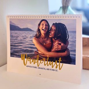 A calendar propped on a white table with a photo of two women hugging in the water with the text on the front 'Wanderlust Adventure Awaits, Go Find It'